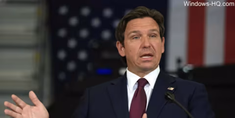 A man in a suit stands at a podium during a speech, with an American flag visible behind him.