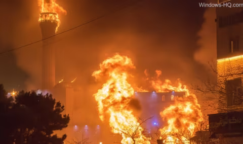 Flames burn near a tower structure at night, surrounded by smoke and trees.