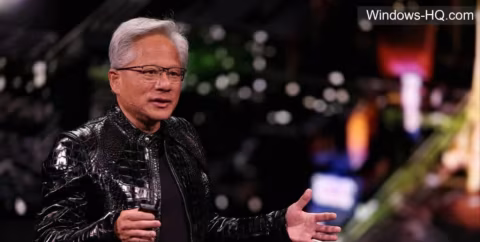 A man wearing glasses and a shiny black leather jacket speaks while sitting in a conference setting.