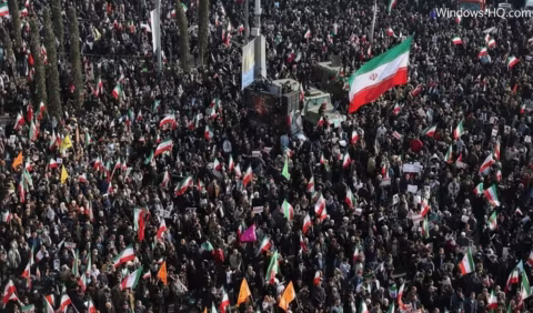 Large crowd of people carrying flags, including Iranian flags, gathered in an outdoor area.