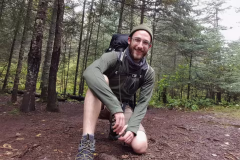A man in a green jacket and knit hat crouches on a muddy trail surrounded by trees.