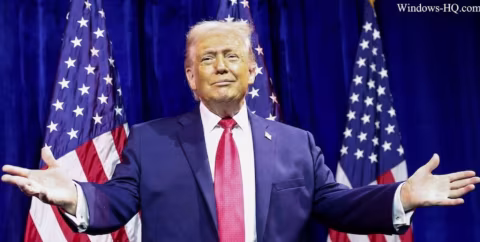 Donald Trump, wearing a suit, standing in front of two American flags.