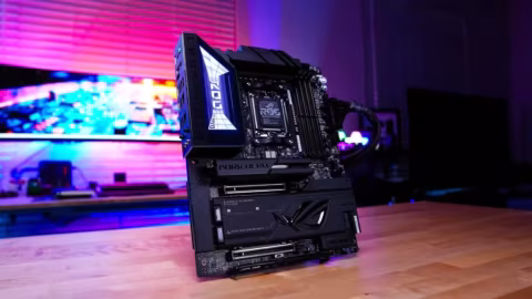 A dark colored computer motherboard with attached graphics card, positioned on a wooden desk.