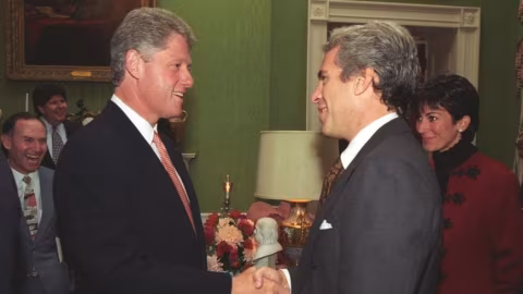 Two men in suits shake hands indoors, flanked by several other people.