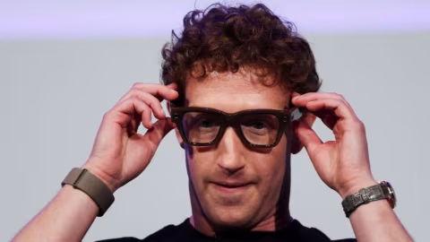 A man with curly brown hair wearing square, dark-rimmed glasses, adjusting the glasses with both hands.