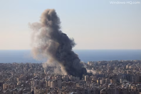 Large plume of smoke rises above an urban cityscape under a clear sky.