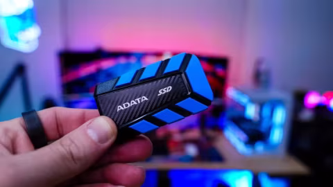 A hand holds a blue and black portable SSD drive in front of a blurred background of digital electronics.