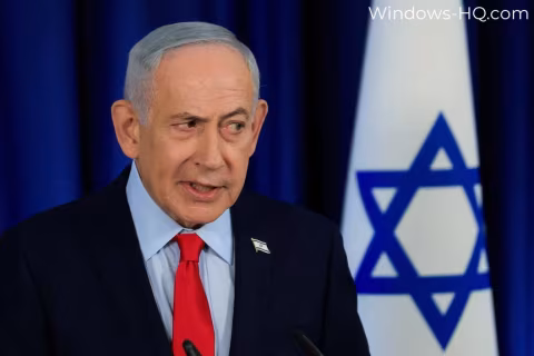 Man in a dark suit speaking at a podium next to an Israeli flag.