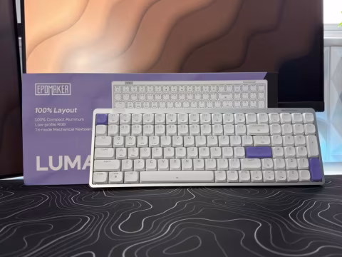 A 100% layout keyboard placed on a dark patterned surface, next to a computer screen.