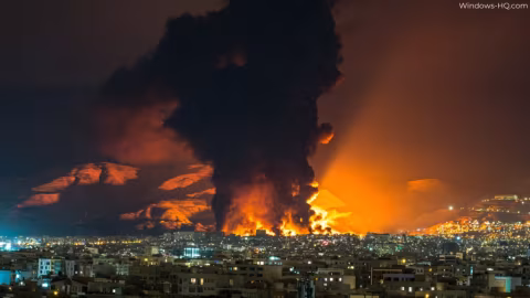 Large column of black smoke rises above a city skyline next to a bright orange fire.