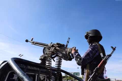 A man wearing a helmet stands beside a mounted automatic weapon in an open vehicle against a blue sky.