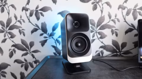 A silver and black computer speaker placed on a dark surface against a patterned wallpaper.