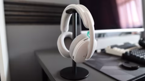 Wireless over-the-ear headphones displayed on a black stand next to a computer monitor.