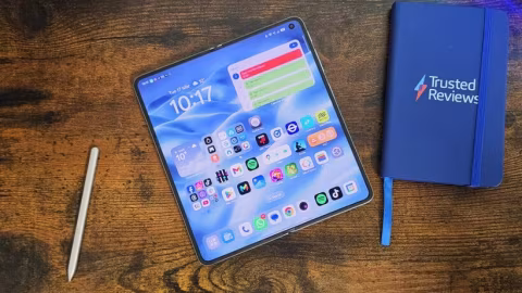 A smartphone with a colorful home screen, resting on a wooden surface next to a blue notebook.