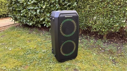 Portable black speaker unit with illuminated circular speaker grilles placed on grass in front of dense green shrubbery.