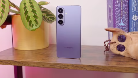 A purple Samsung smartphone with a triple-lens camera setup sits on a wooden shelf next to a wooden skull and a green potted plant.