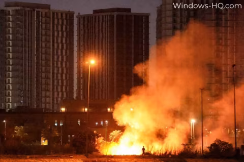 Large column of orange smoke rising behind burning debris near tall buildings.