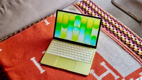A light yellow MacBook laptop with a colorful rainbow keyboard rests on a patterned textile surface.