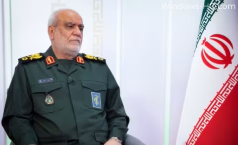 A man in military uniform sits next to a large flag featuring the Iranian emblem.