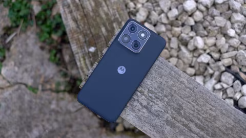 A dark smartphone lying on a wooden plank, viewed outdoors near rocks and foliage.