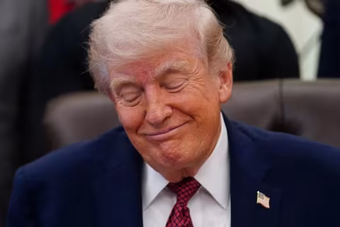 A man with blonde hair wearing a navy suit and red patterned tie smiles while looking down.