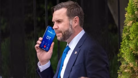 Man in suit holds smartphone displaying 'Donald J. 1 DT' text.
