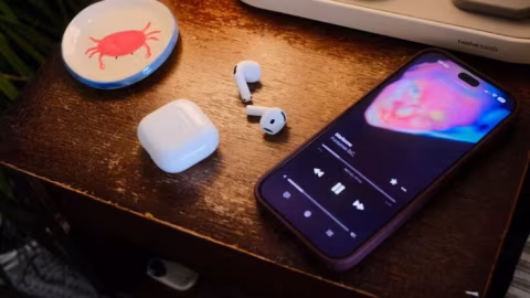White wireless earbuds and charging case are placed on a dark wood surface next to a smartphone showing a music player interface.