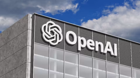 The OpenAI logo and name displayed on the upper facade of a modern building against a blue, cloudy sky.