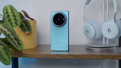 Light blue Vivo smartphone, white wireless headphones on a stand, and green plant foliage on a wooden surface.