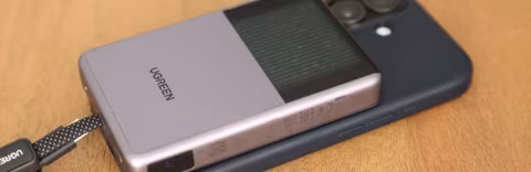 A silver rectangular portable charger branded UGREEN rests on a wooden surface next to a dark smartphone.