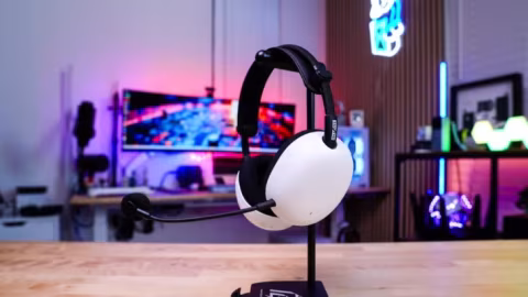 White and black over-the-ear headset displayed on a stand, placed on a wooden table in front of a brightly lit monitor.
