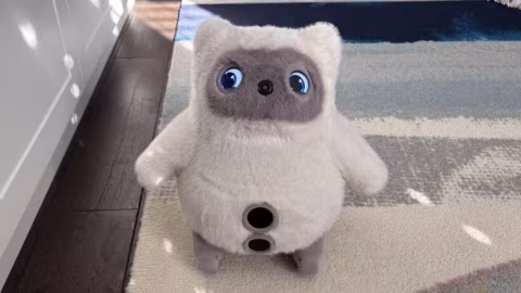 A plush gray robot toy with blue eyes sits on a patterned rug near a dark wooden floor.