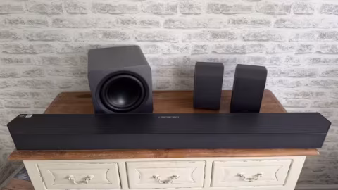 Black speaker components, including a long soundbar and multiple vertical speakers, displayed on a wooden dresser against a textured wall.