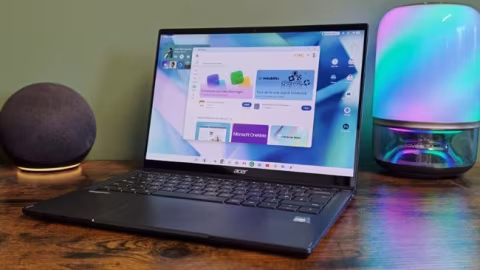 Open laptop displaying a Google interface next to a cylindrical lamp with multi-colored LED lights on a wooden table.