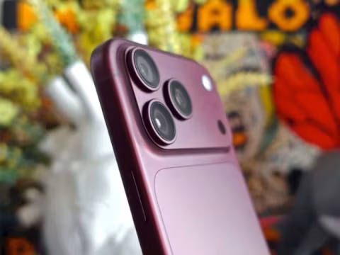 Pink smartphone with a visible triple-lens camera module, positioned in front of a highly blurred background.