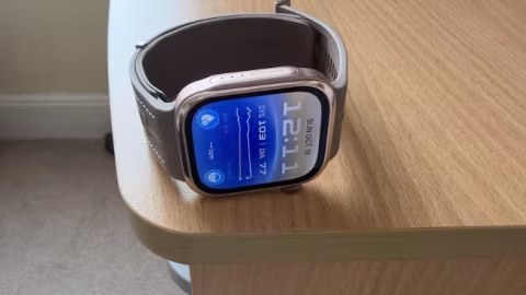 A smartwatch with a dark strap and silver bezel sits on a light wood grain surface. The screen displays 12:21 and date information.