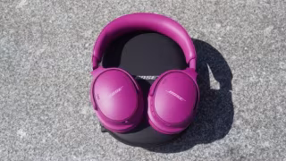 A pair of magenta, over-ear headphones with "Bose" visible on the earcups, resting on a black pad on gray concrete.
